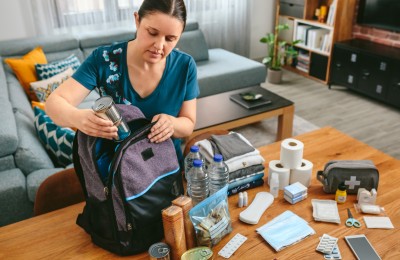 Woman putting cans of food to prepare emergency backpack in living room