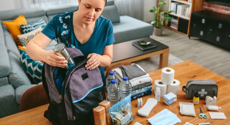 Woman putting cans of food to prepare emergency backpack in living room